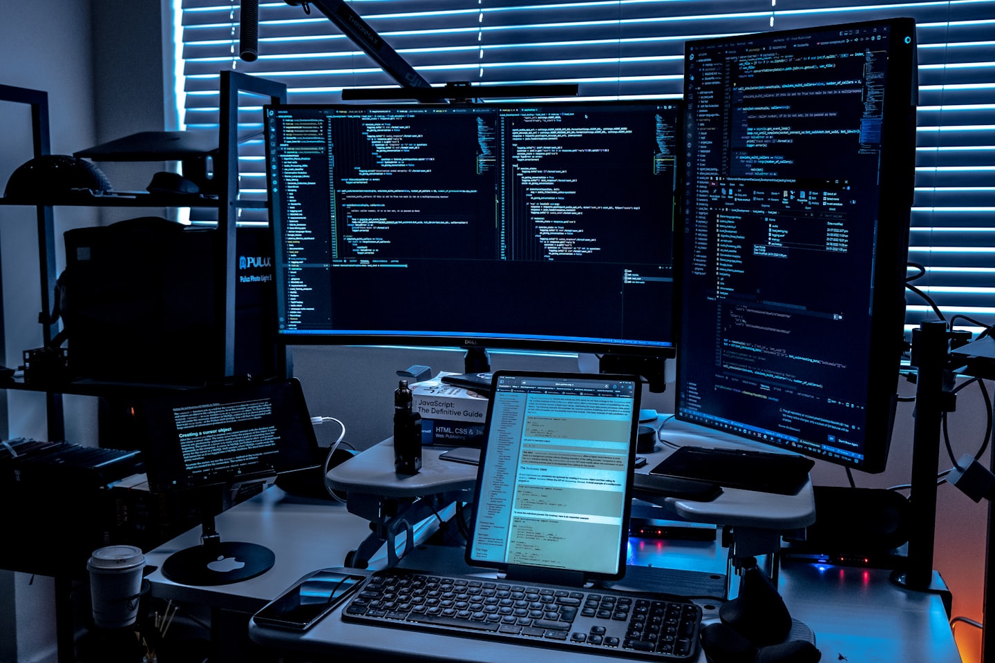 Dos monitores grandes y un portátil mostrando código en un espacio de trabajo oscuro.